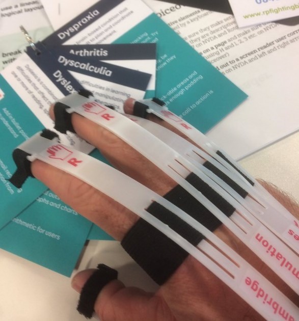 photo of a hand wearing simulation gloves, together with cards describing dyslexia, dyspraxia and arthritis